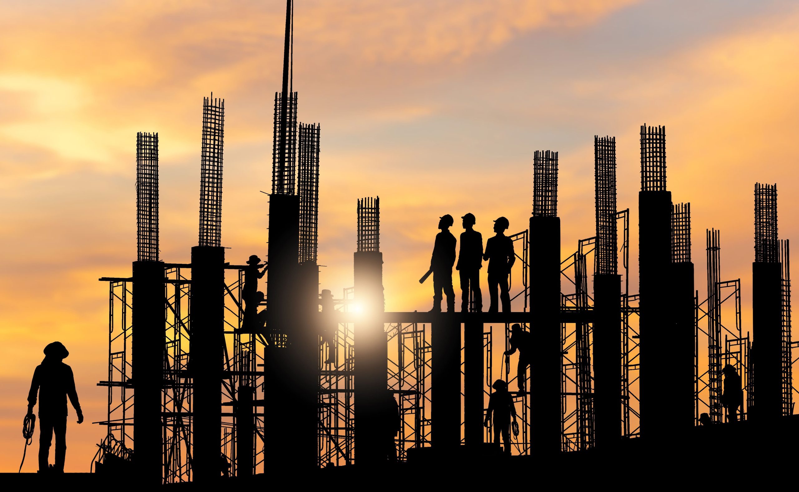 Silhouettes of construction workers and steel framework against an orange sunset sky